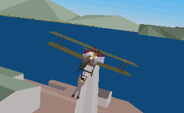 A screenshot of Stunt Island for DOS. A stuntman wearing grey overalls hangs precariously from the undercarriage of a light aircraft, as it flies over a group of buildings. 